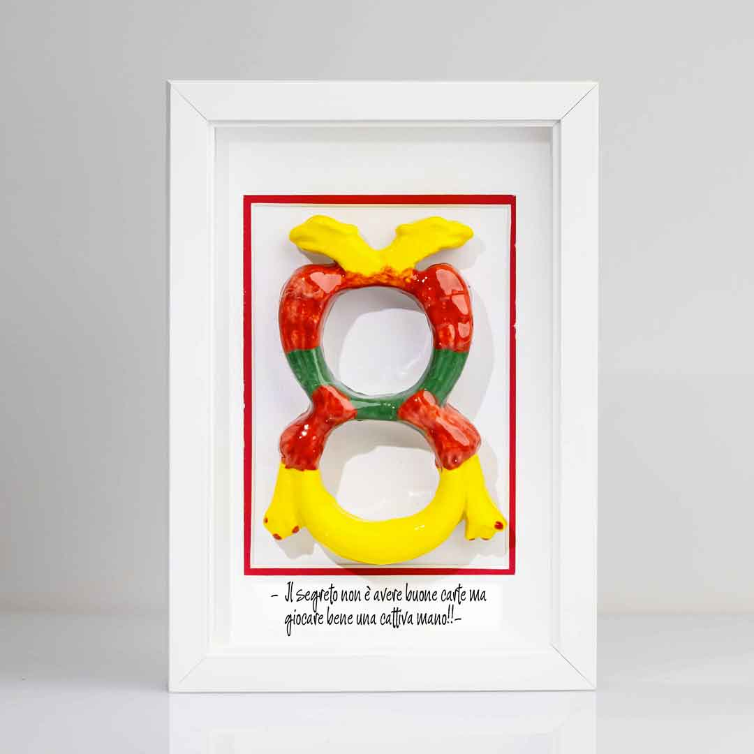 Quadro con contenente una riproduzione tridimensionale in ceramica dell'Asso di Denari delle carte napoletane, raffigurato come una grande moneta o sole stilizzato con decorazioni geometriche e antropomorfe nei toni del giallo, rosso e verde. In basso è riportata la scritta in corsivo: "Il segreto non è avere buone carte ma giocare bene una cattiva mano!!". Cornice tutta bianca