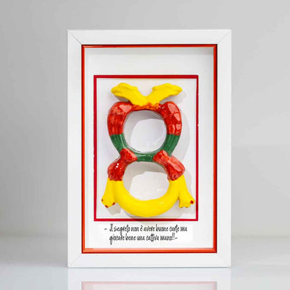 Quadro contenente una riproduzione tridimensionale in ceramica dell'Asso di Denari delle carte napoletane, raffigurato come una grande moneta o sole stilizzato con decorazioni geometriche e antropomorfe nei toni del giallo, rosso e verde. In basso è riportata la scritta in corsivo: "Il segreto non è avere buone carte ma giocare bene una cattiva mano!!". Il bordo interno della cornice è rosso