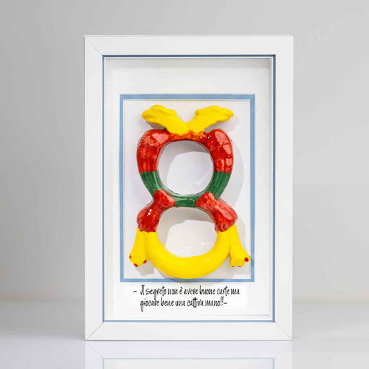 Quadro con cornice contenente una riproduzione tridimensionale in ceramica dell'Asso di Denari delle carte napoletane, raffigurato come una grande moneta o sole stilizzato con decorazioni geometriche e antropomorfe nei toni del giallo, rosso e verde. In basso è riportata la scritta in corsivo: "Il segreto non è avere buone carte ma giocare bene una cattiva mano!!". Il bordo interno della cornice è azzurro